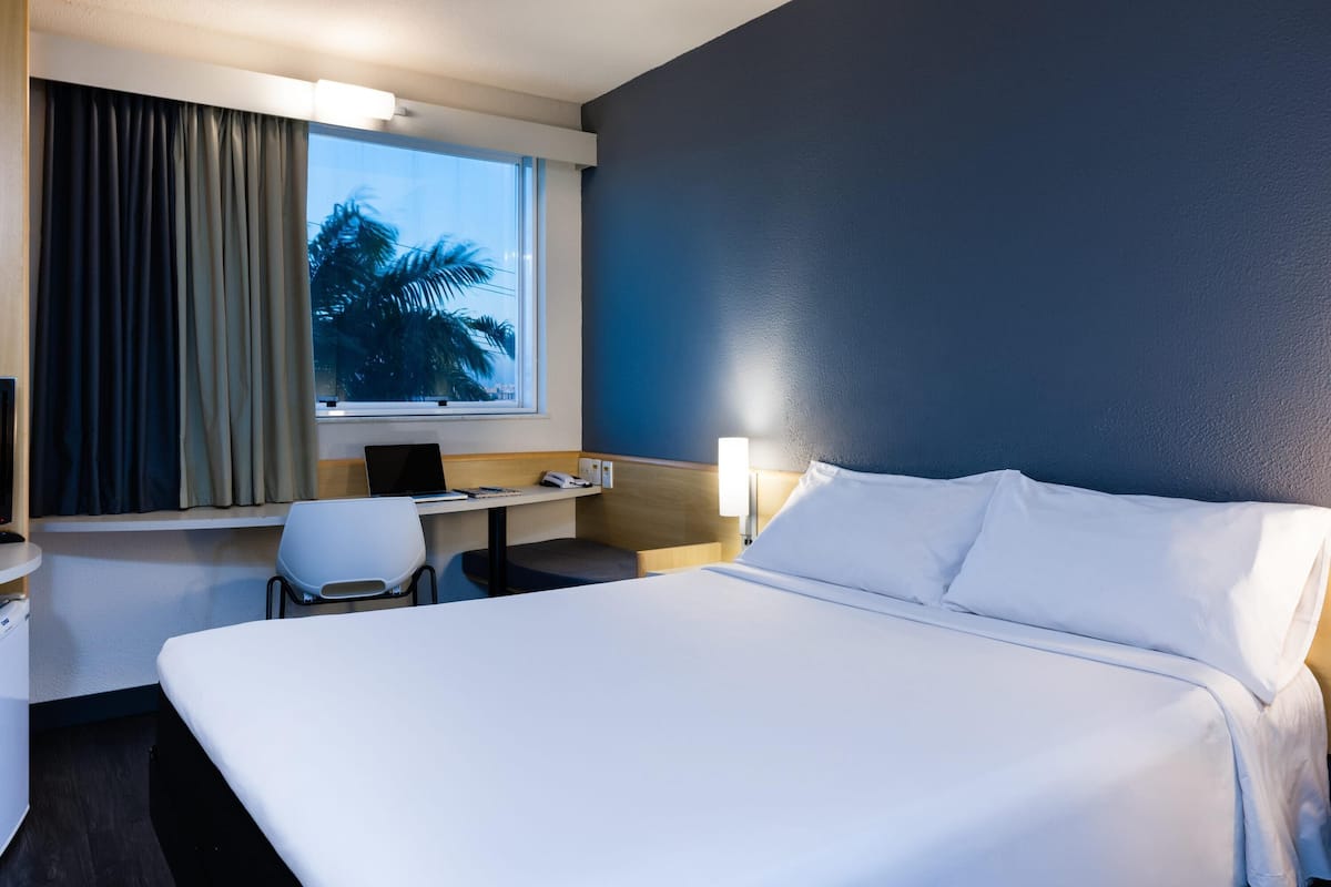 A hotel room with a large bed, a desk with a laptop, a chair, and a window with curtains.