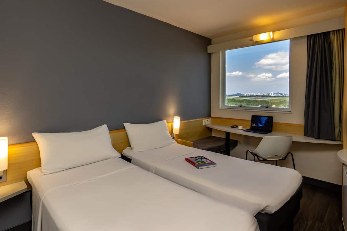 A hotel room with two beds, a desk with a laptop, a chair, and a window with a view of a cityscape.
