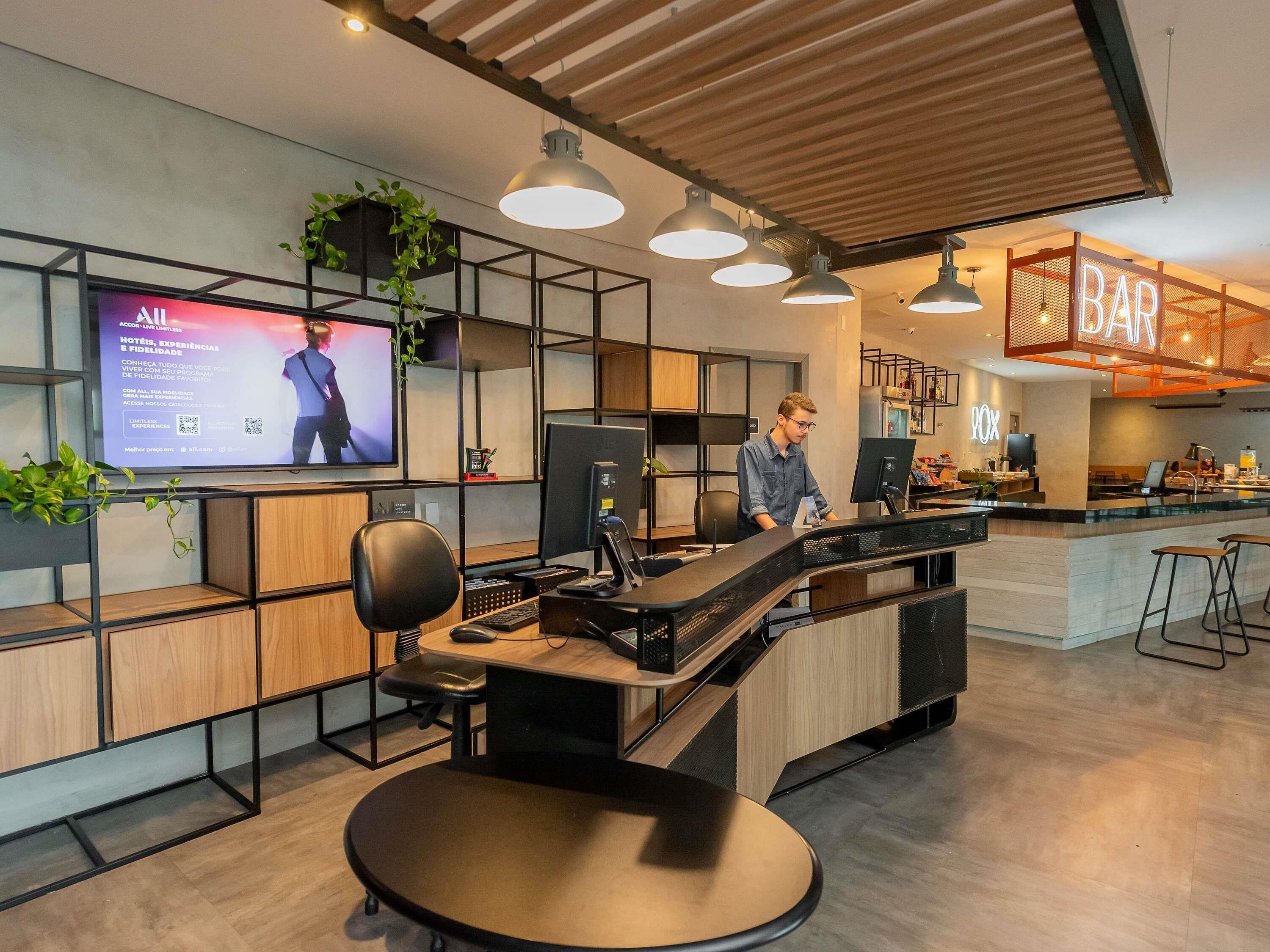 Zona de recepción moderna de hotel con una pantalla digital mostrando información, un mostrador con una computadora y un área de bar al fondo.