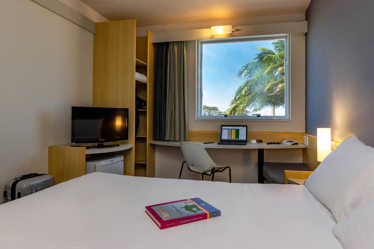 Ein Hotelzimmer mit Bett, Fernseher, Schreibtisch mit Laptop, Stuhl und Fenster mit Blick auf Palmen.