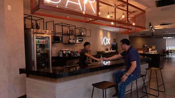 Zona de bar con un letrero de neón, un refrigerador lleno de bebidas y una barra donde dos personas interactúan.