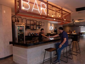 Un espace bar agrémenté d’un néon, d’un réfrigérateur rempli de boissons et d’un comptoir où deux personnes sont en train de discuter.