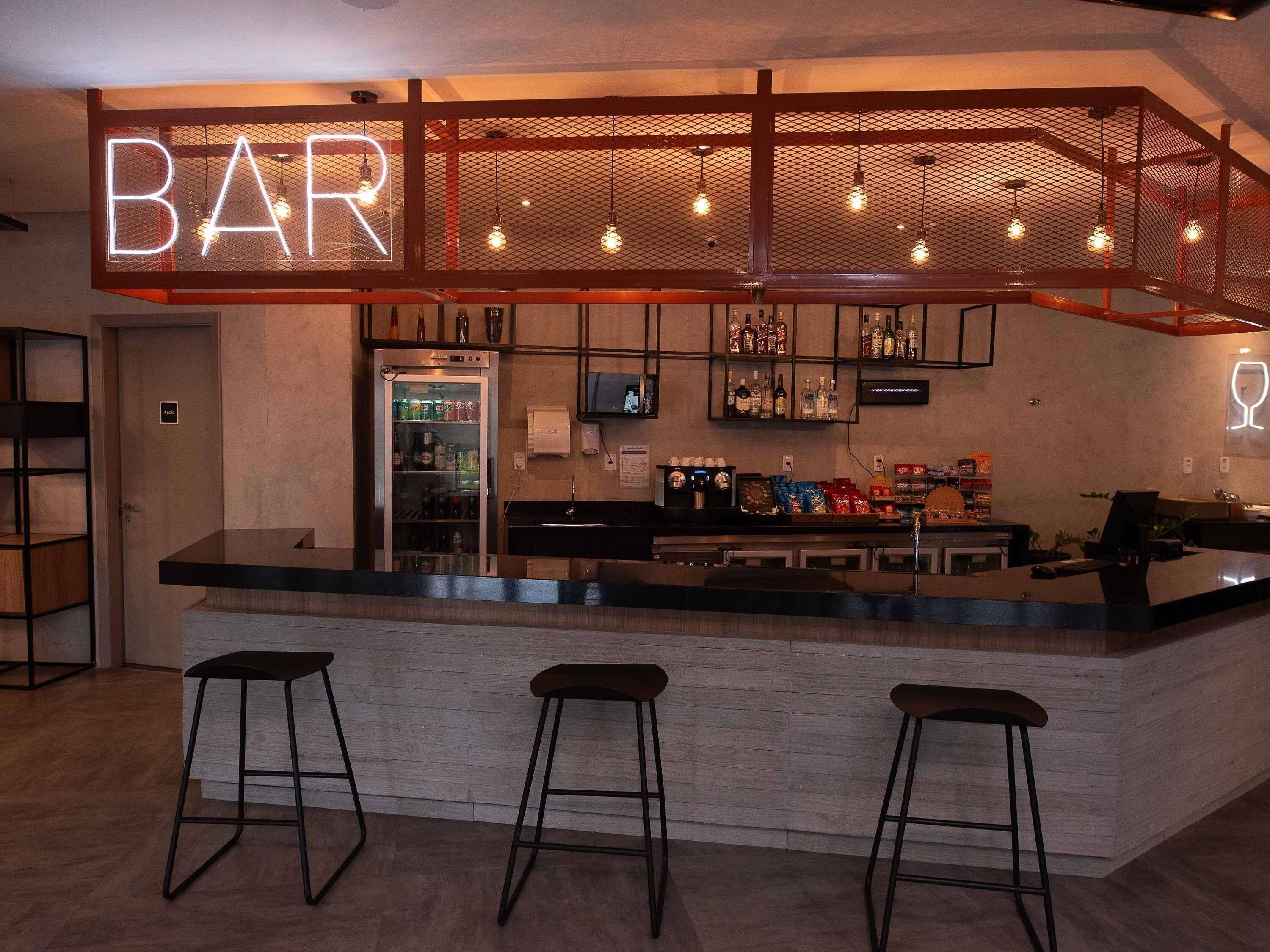 Un espace bar agrémenté d’un néon, d’un réfrigérateur rempli de boissons et d’un comptoir avec des tabourets.