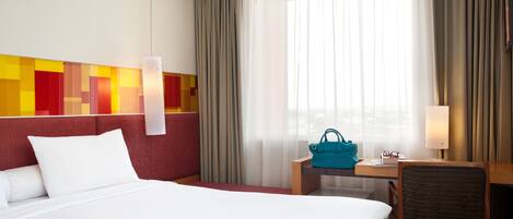 Habitación de hotel con una cama grande, un escritorio con silla, un bolso color turquesa y una ventana con cortinas.