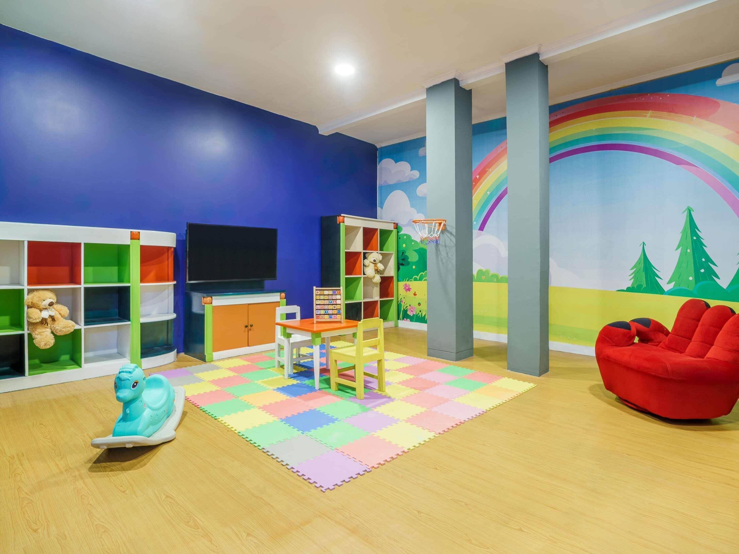 A children's playroom with a blue wall, a rainbow mural, a TV, a bookshelf, a rocking horse, and a red chair.