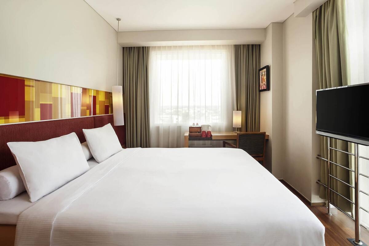 A hotel room with a large bed, a TV, a desk with a lamp, and a window with curtains.