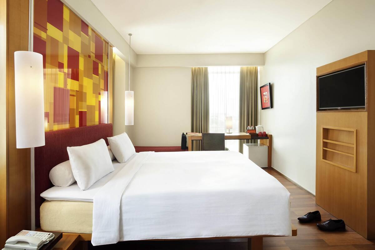 A hotel room with a bed, a desk, a chair, a TV, and a shoe rack.