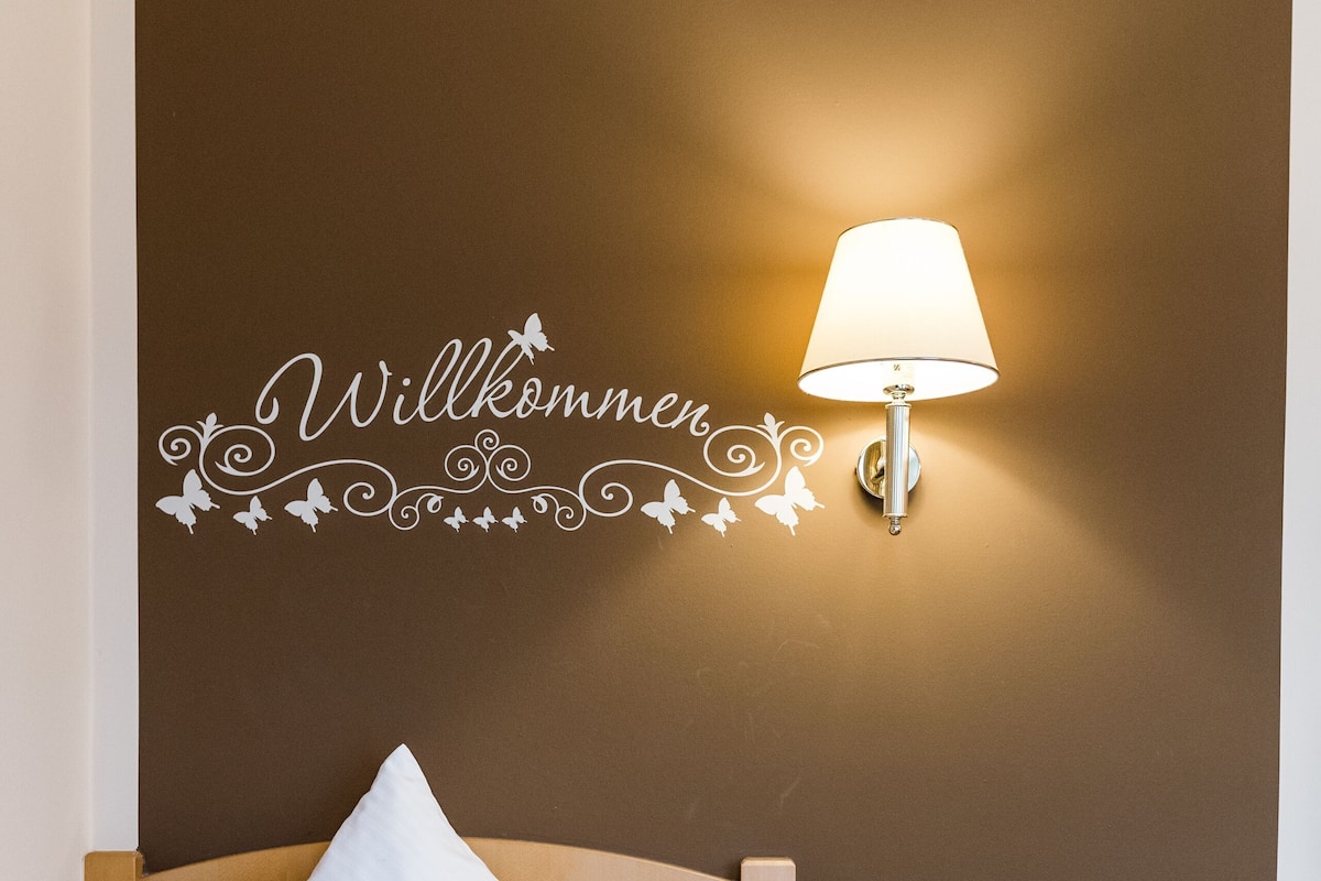 Une chambre d’hôtel avec un lit, une table de chevet, une lampe, un panier de fruits et un panneau d’accueil sur le mur.
