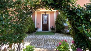 A pink building with a cobblestone pathway leading to a door. There are potted plants and a small archway of greenery in front. - Vila Duparchy (Mealhada)