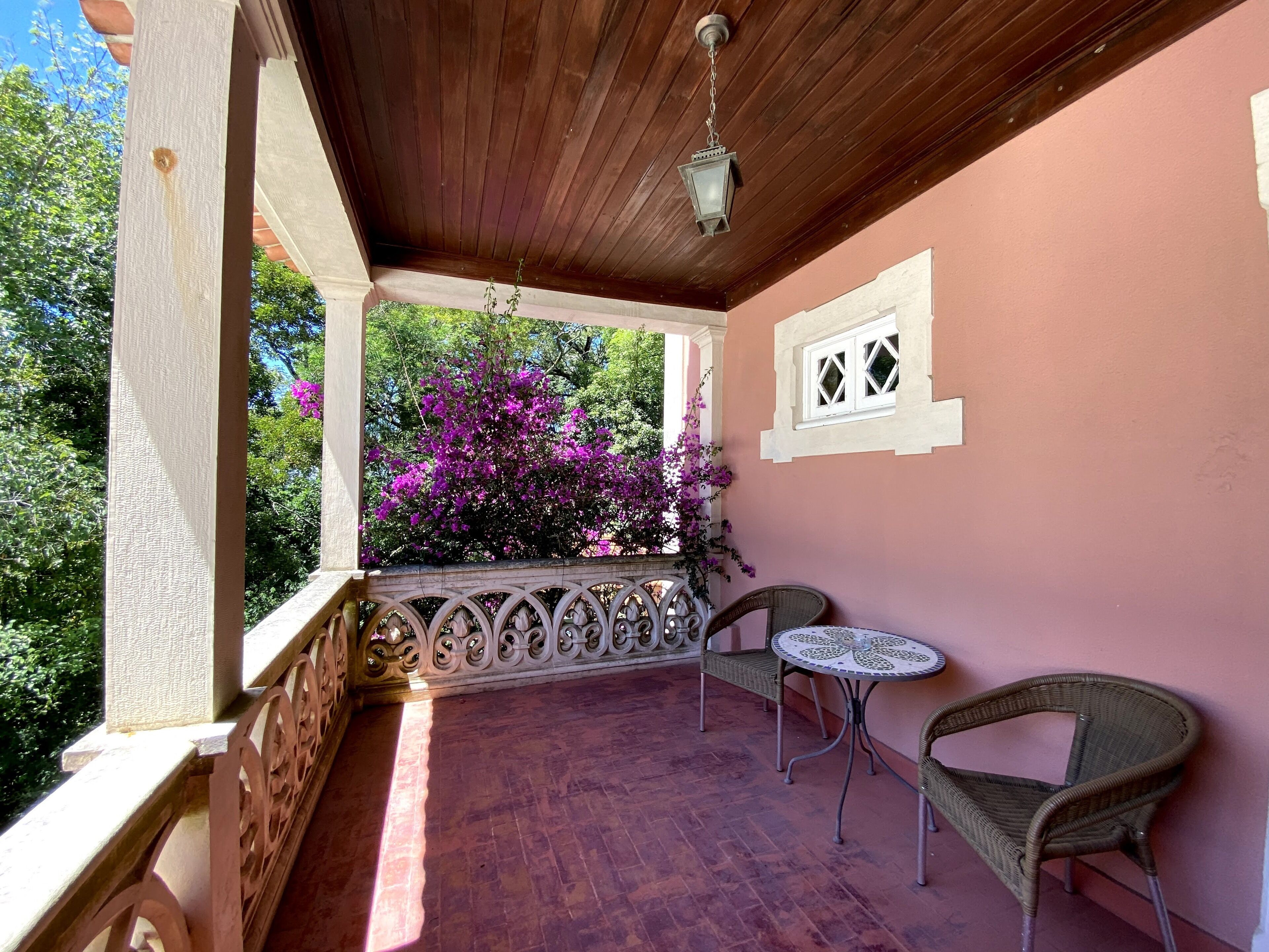 A covered outdoor area with a wooden ceiling, a white railing, and a view of purple flowers. There are two wicker chairs and a small round table.