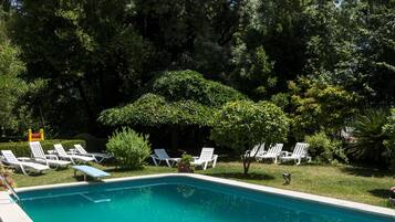 Piscina rectangular con agua azul cristalina, rodeada de sillones blancos y follaje.