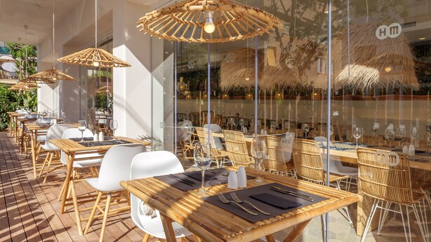 An outdoor dining area with wooden tables and chairs, bamboo umbrellas, and glass walls reflecting the interior.