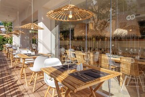 Un área de comedor al aire libre con mesas y sillas de madera, sombrillas de bambú y paredes de vidrio que reflejan el interior.