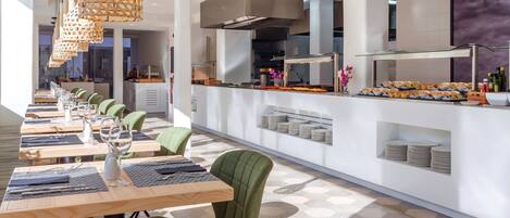 A modern dining area with long tables, green upholstered chairs, and a central island with food displays.