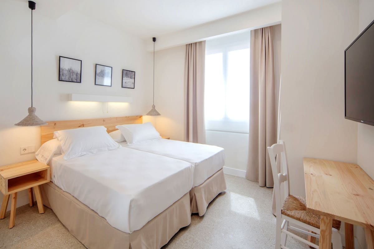 A hotel room with two beds, a wooden nightstand, a desk, and a chair. There are three framed pictures on the wall, a window with curtains, and a flat-screen TV mounted on the wall.