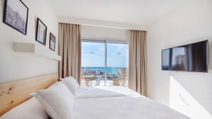 A hotel room with a large bed, a TV mounted on the wall, and a balcony with a view of the sea.