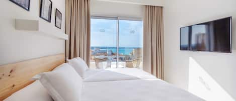 A hotel room with a large bed, a TV mounted on the wall, and a balcony with a view of the sea.