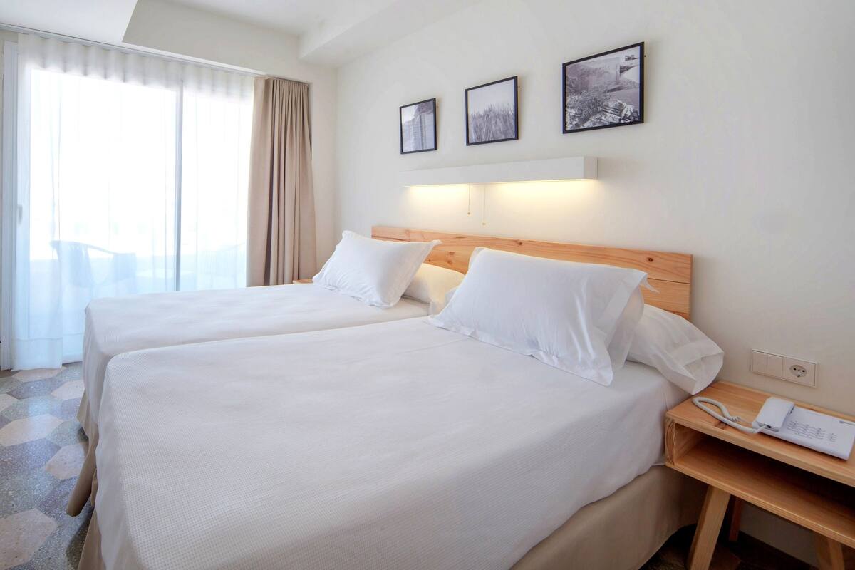 A hotel room with two beds, a wooden headboard, a bedside table with a phone, and a large window with curtains.