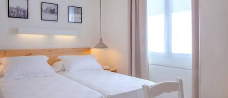 A room with two single beds, a wooden headboard, a hanging light fixture, and a window with curtains.