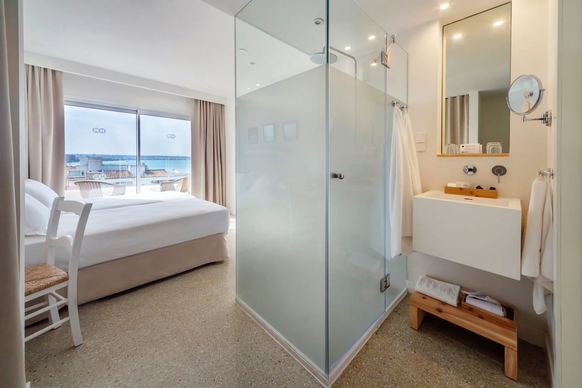 A hotel room with a bed, a glass-enclosed shower, a sink, and a view of the sea.