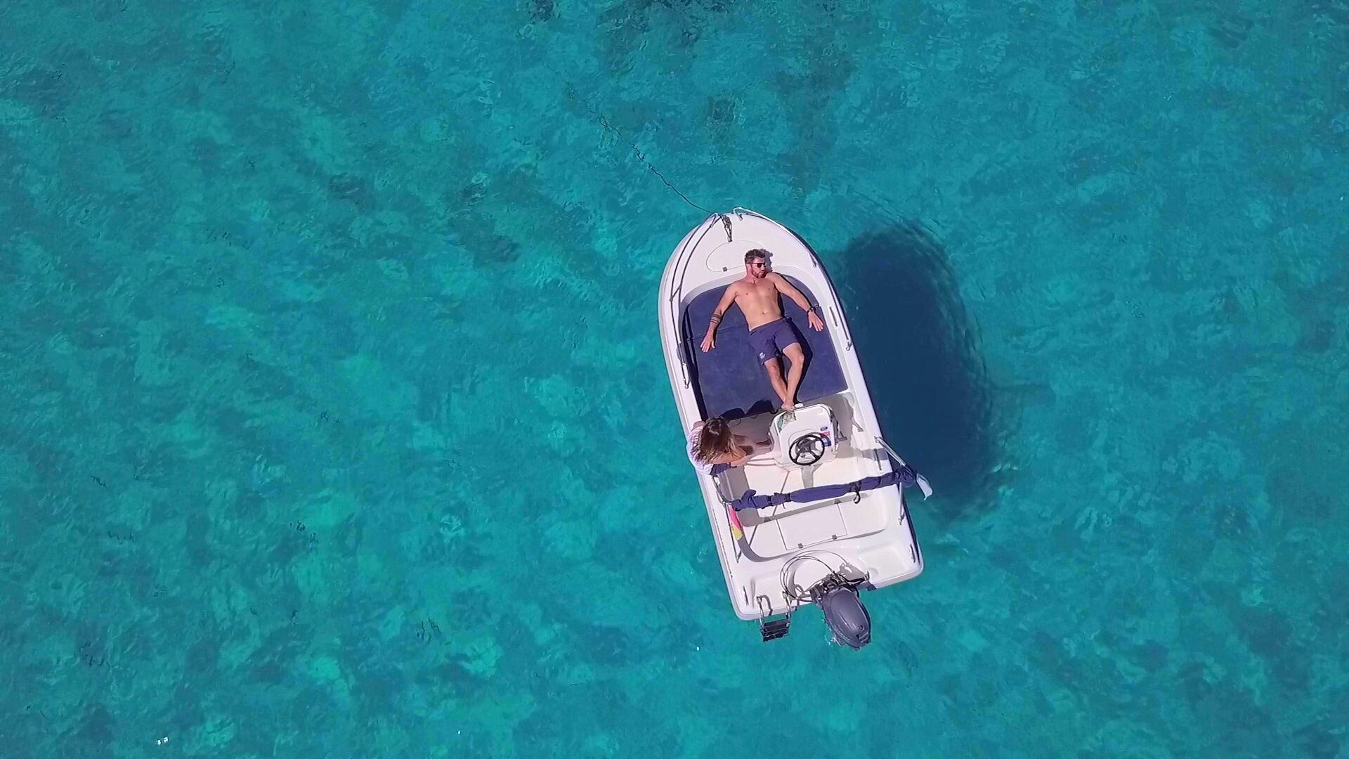 An aerial view of a small boat with two people on it, floating on clear turquoise water.