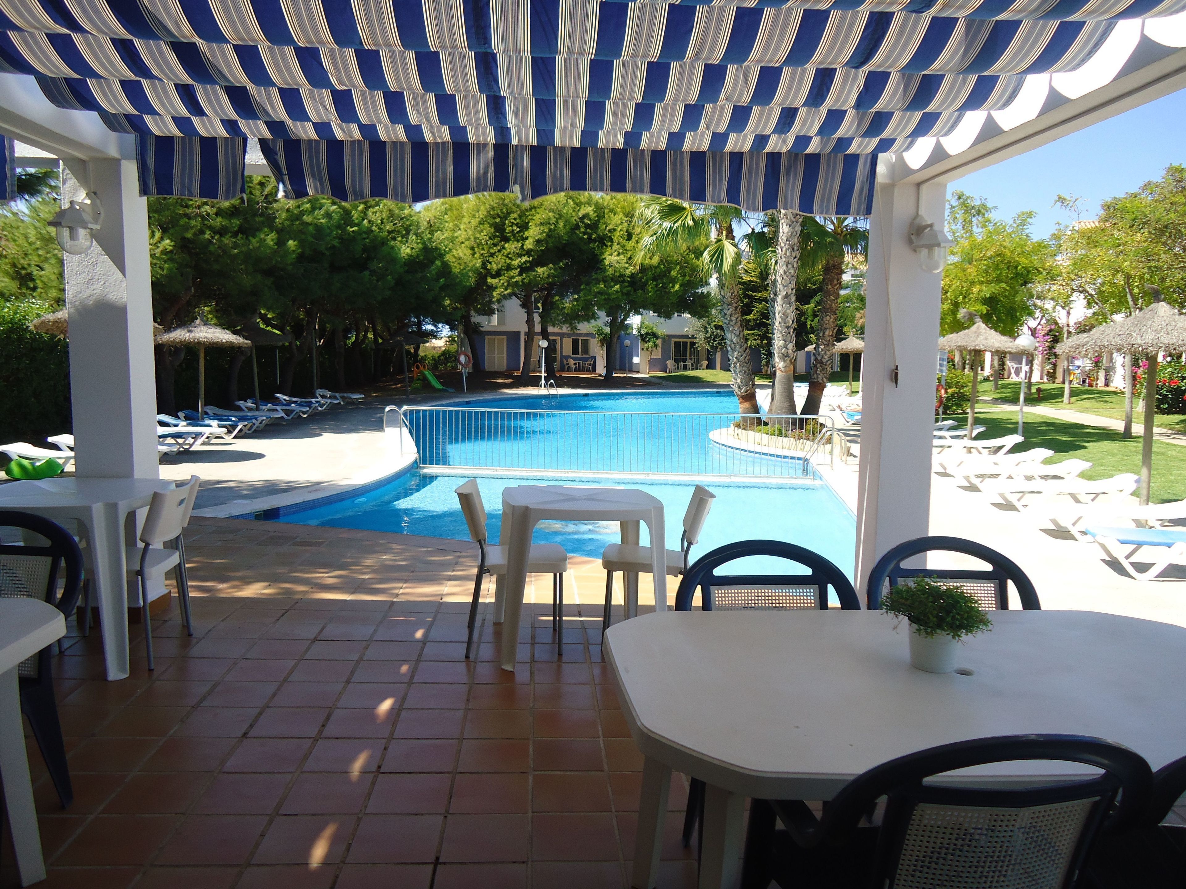 A pool area with a tiled floor, lounge chairs, and a covered seating area with a striped awning.