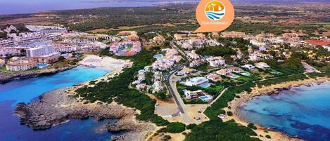 Vista aérea de un pueblo costero con un puerto deportivo, propiedades frente a la playa y el logotipo de Club Ciudadela.