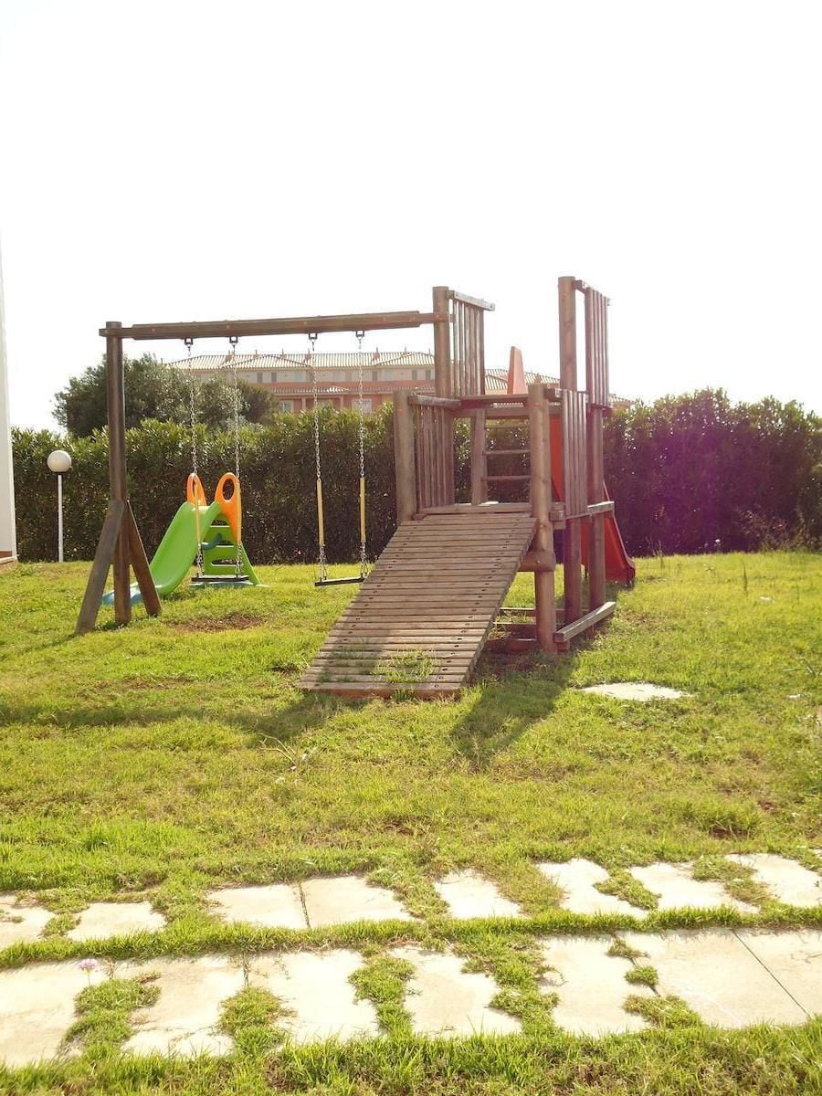 Une structure de jeux en bois, dotée d’un toboggan et de balançoires, installée sur une pelouse verdoyante, avec un chemin pavé.