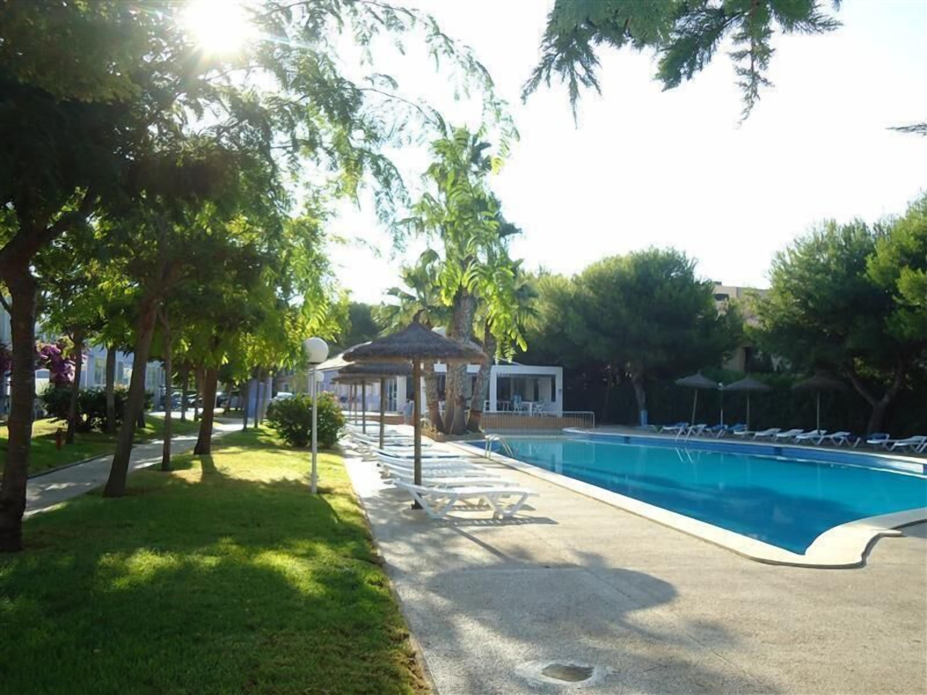 Una piscina con sillones de descanso, un pabellón y árboles que la rodean.