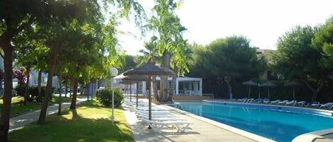 Una piscina con sillones de descanso, un pabellón y árboles que la rodean.