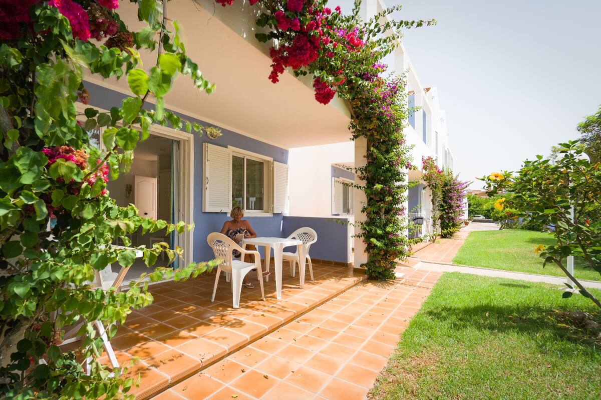 A patio with white chairs and a table, surrounded by a garden with flowering plants.
