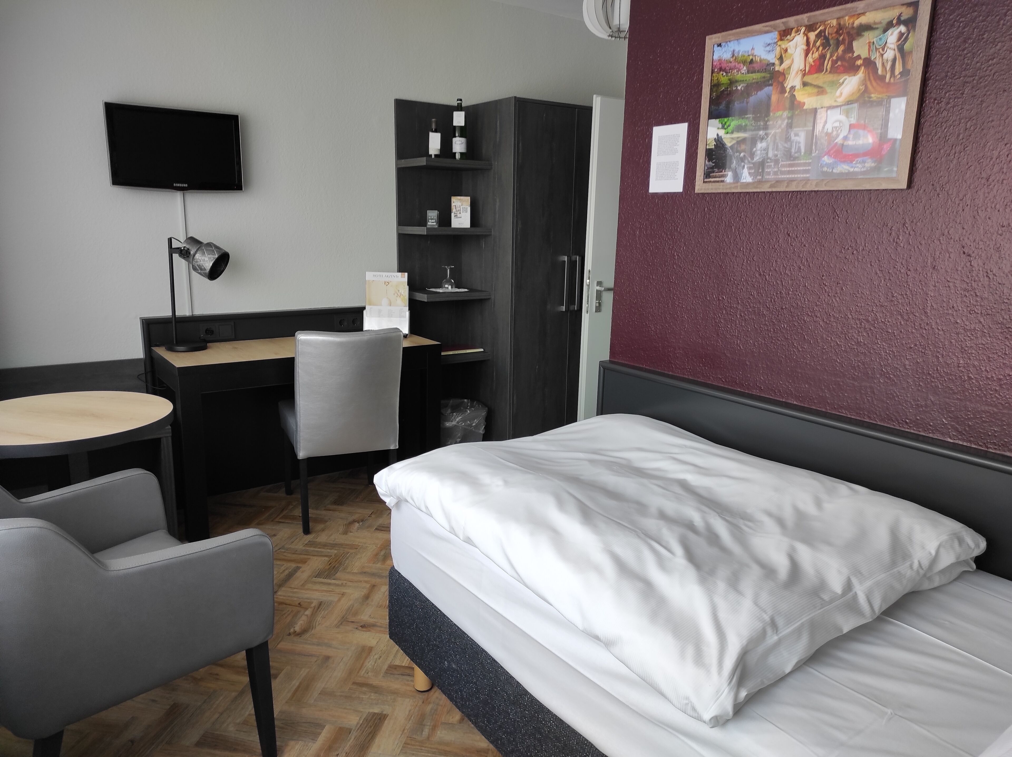 Habitación de hotel con cama, escritorio, silla, televisor y estante en la pared con objetos.