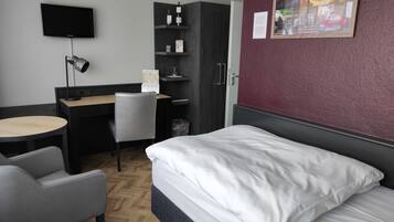 Habitación de hotel con cama, escritorio, silla, televisor y estante en la pared con objetos.