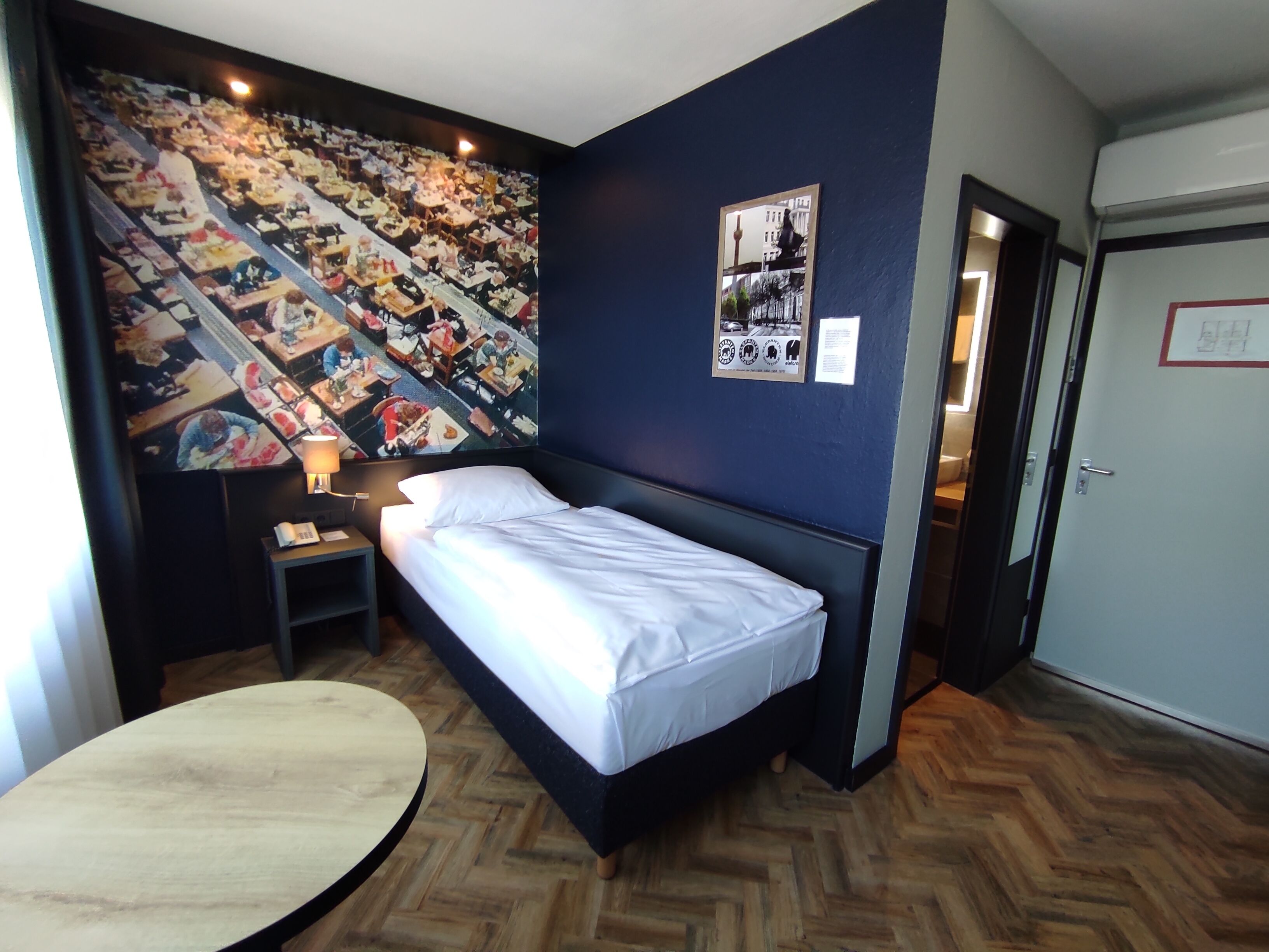 A hotel room with a bed, a round table, a nightstand, a wall mural, and a door.