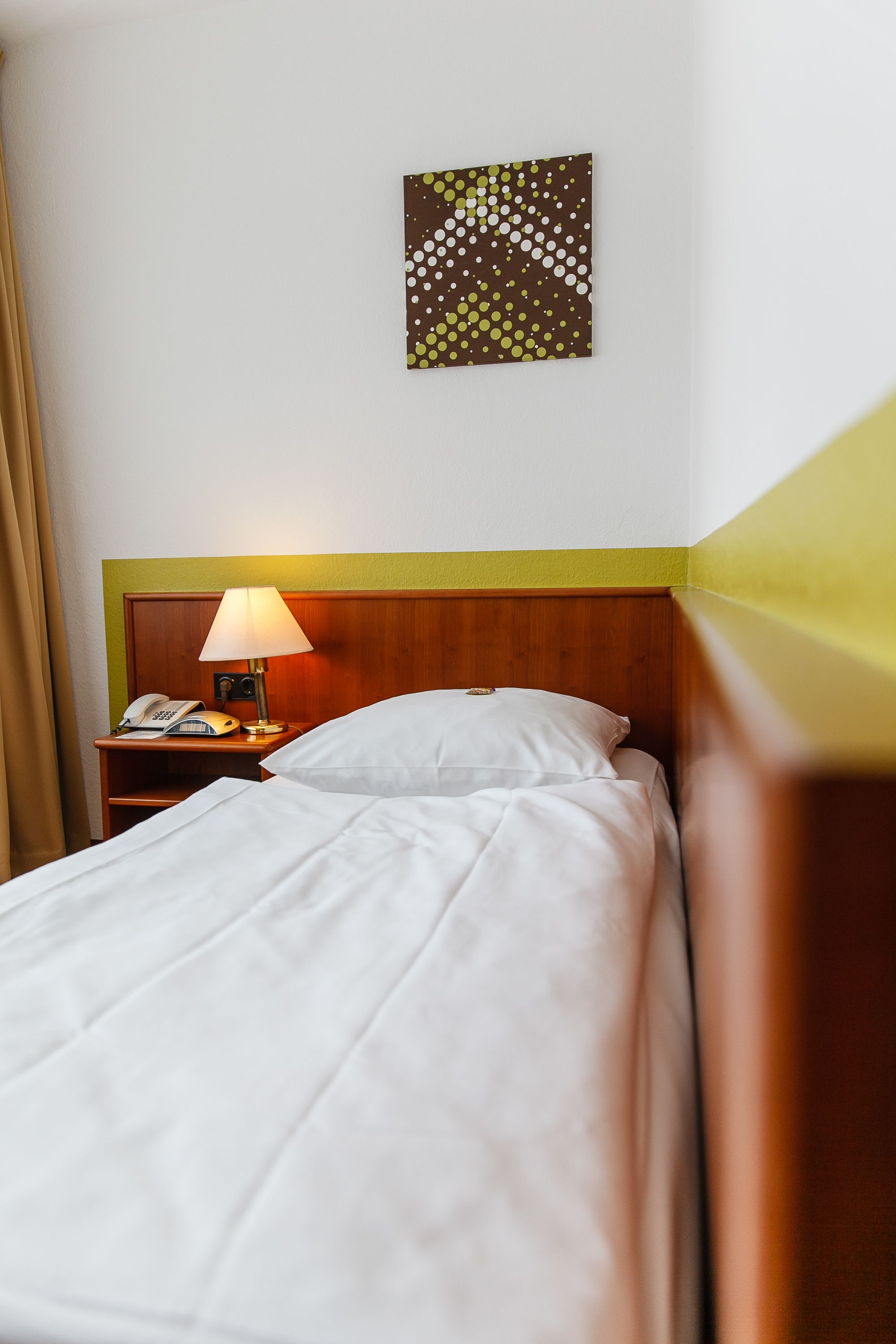 A hotel room with a wooden headboard, a bedside table with a lamp, a telephone, and a wall-mounted artwork.