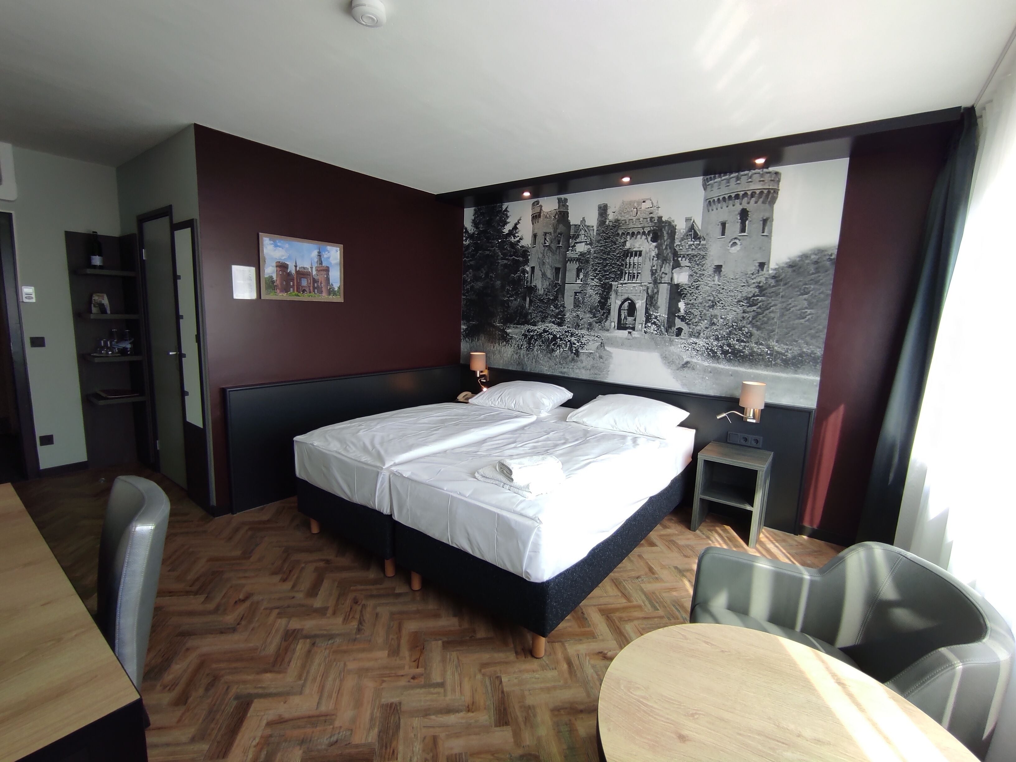 A hotel room with a large bed, a desk, and a chair. There is a painting on the wall and a window with a view of a castle.