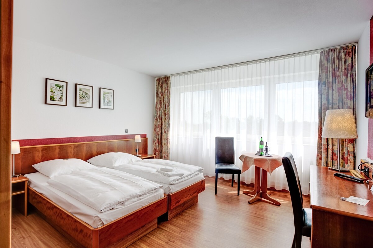 A hotel room with a double bed, a desk with a chair, a small table, and a window with sheer curtains.