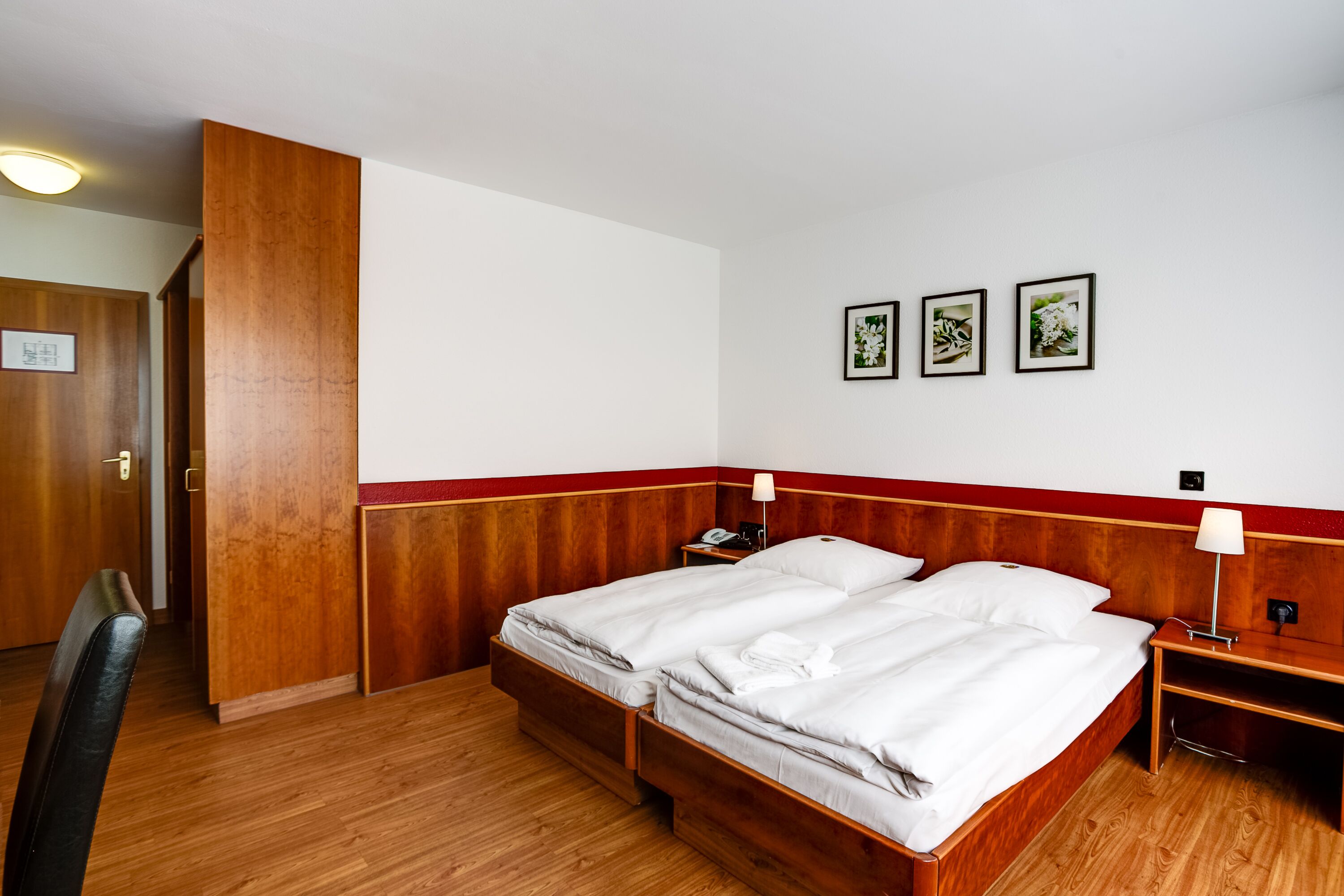 Ein Hotelzimmer mit einem hölzernen Bett, einem Schreibtisch und einem Stuhl. An der Wand hängen drei gerahmte Bilder.