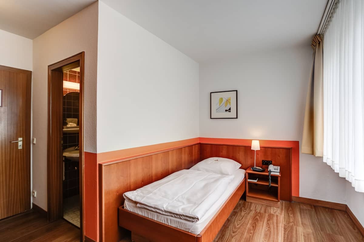 Ein Hotelzimmer mit einem hölzernen Bett, einem Nachttisch mit Lampe, einem gerahmten Bild an der Wand und einem Fenster mit Vorhängen.