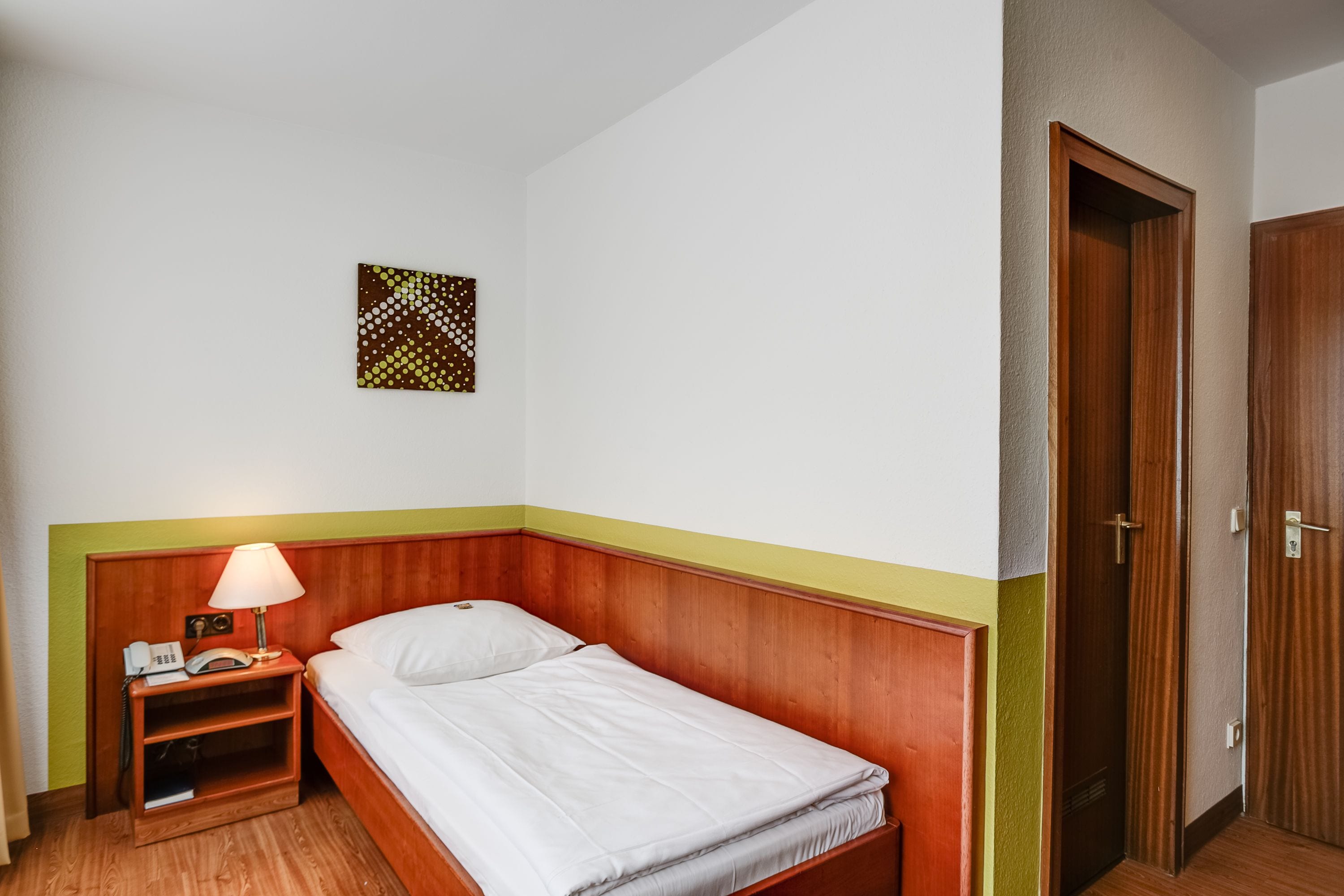 Una cama individual con cabecera de madera, una mesita de noche con lámpara, y una pequeña obra de arte en la pared.