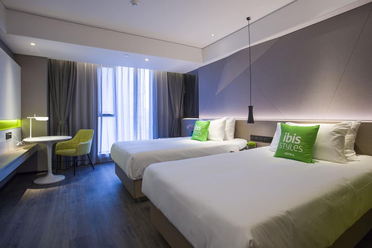 Quarto de hotel moderno com duas camas, uma escrivaninha, uma cadeira e uma janela grande com cortinas.