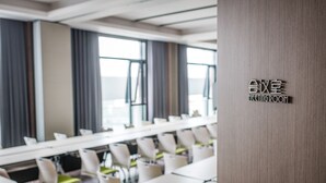 Une salle de réunion moderne, avec des rangées de tables et de chaises, de grandes fenêtres dotées de rideaux et un panneau indiquant qu’il s’agit d’une salle de réunion.
