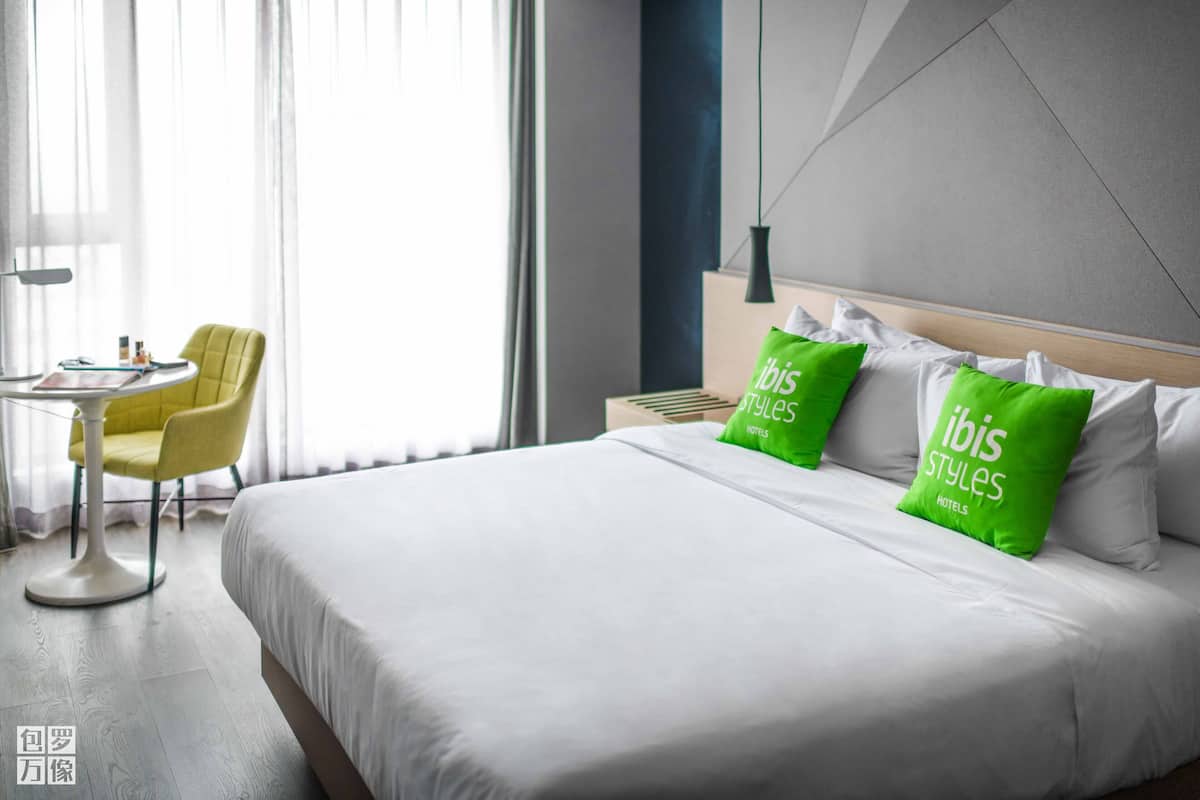 Quarto de hotel com uma cama grande, duas almofadas verdes, uma cadeira amarela, uma mesinha e uma janela com cortinas leves.