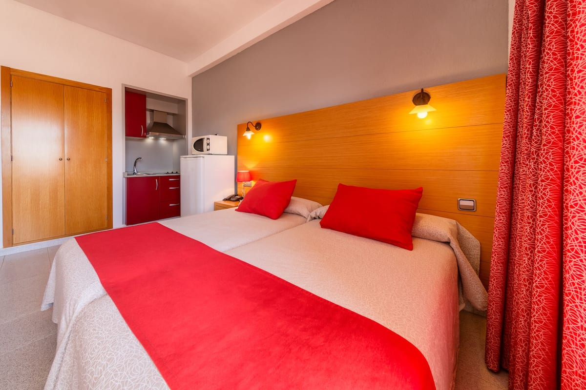 Quarto de hotel com uma cama grande, roupa de cama vermelha, cabeceira de madeira, mesa de cabeceira, guarda-roupa vermelho e uma kitchenette ao fundo.