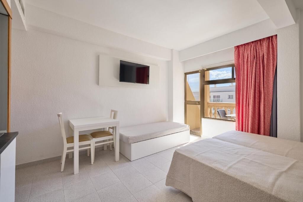 A hotel room with a bed, a desk, a chair, a TV mounted on the wall, and a window with curtains.