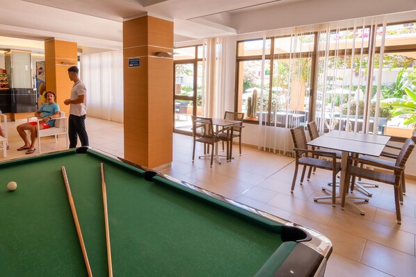 A pool table in a well-lit indoor area with natural light from large windows, a dining area with wicker furniture, and a visible sign indicating a pool table rental service. - Apartamentos Isla del Sol (Palma de Mallorca)