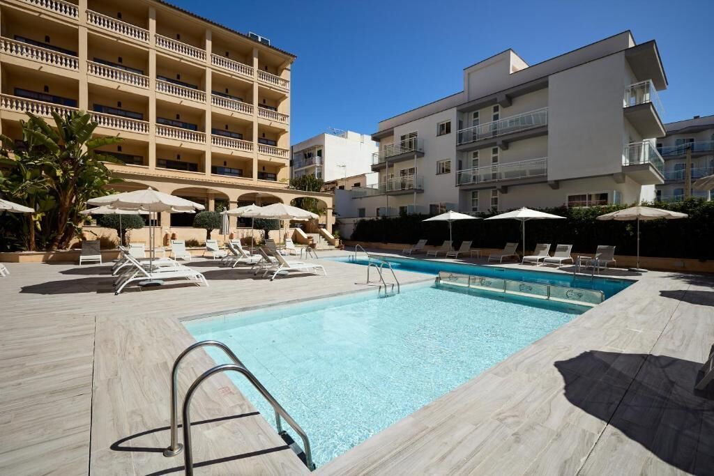 Una piscina con sillones y sombrillas, rodeada de edificios de varios pisos con balcones.