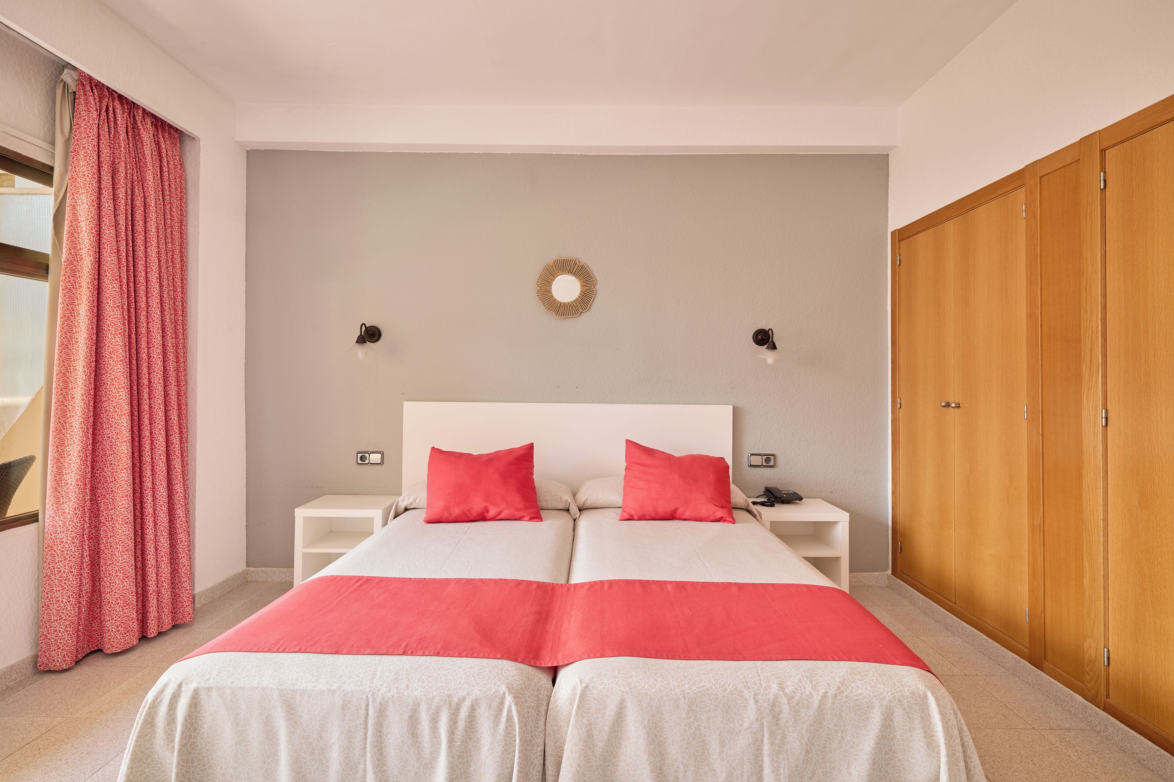Un lit double avec une literie rouge et blanche, deux tables de chevet avec des lampes, une armoire en bois et un luminaire fixé au mur.