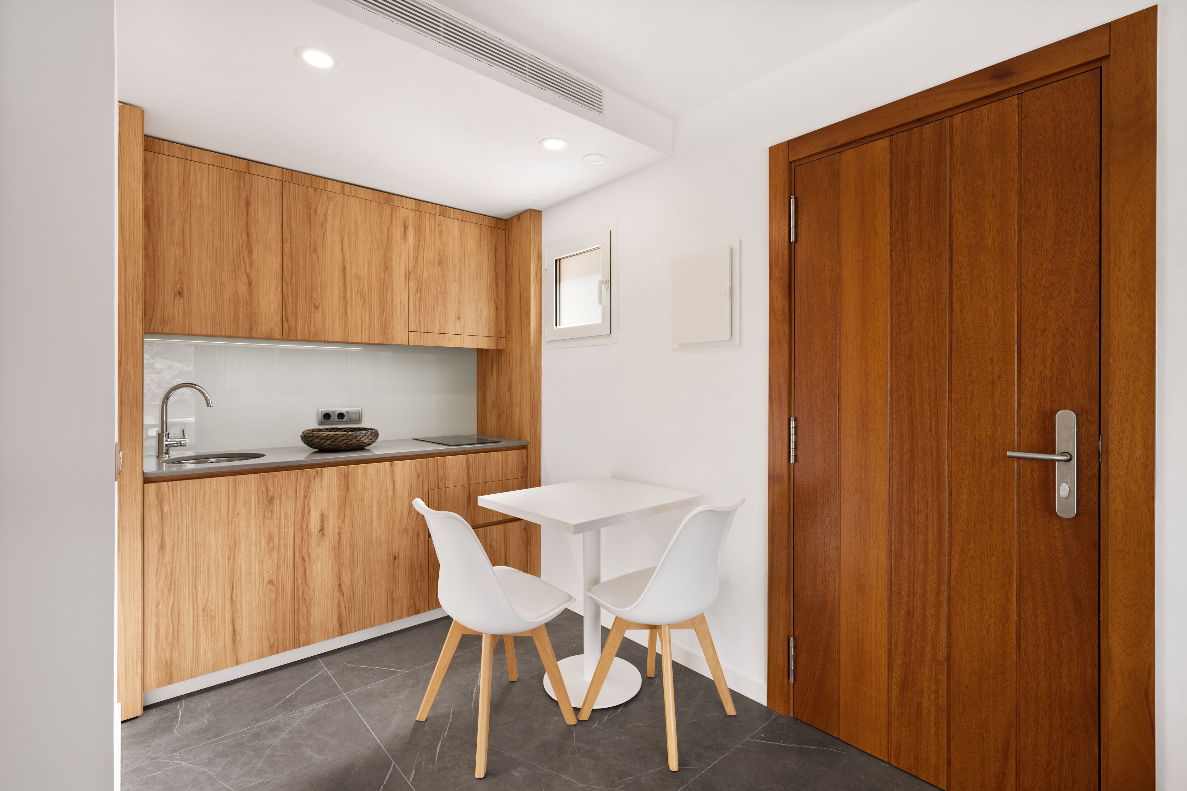 Een kleine keuken met houten kastjes, een wit aanrechtblad en een tafeltje met twee witte stoelen.