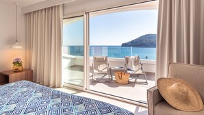 Une chambre d’hôtel avec un grand lit, un balcon donnant sur la mer et une chaise avec un chapeau.